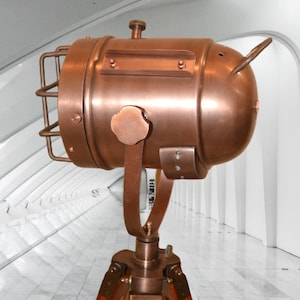 May include: A copper-colored spotlight with a cage around the lens, mounted on a wooden tripod. The spotlight is adjustable and has a vintage industrial design.