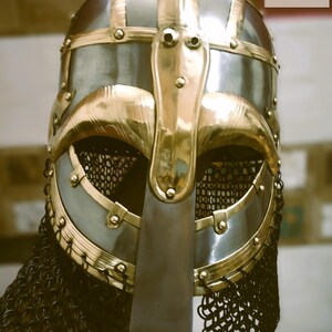 May include: A detailed close-up of a medieval Viking helmet. The helmet features a silver-colored metal base with gold-colored accents and trim. A chainmail neck guard hangs from the helmet. The logo "BHARAT MEDIEVAL DECOR" is in the top right corner.