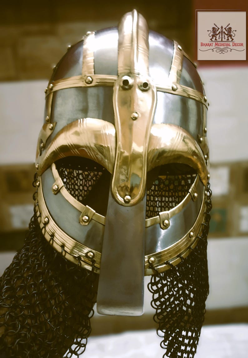 May include: A detailed close-up of a medieval Viking helmet. The helmet features a silver-colored metal base with gold-colored accents and trim. A chainmail neck guard hangs from the helmet. The logo "BHARAT MEDIEVAL DECOR" is in the top right corner.