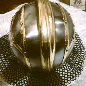 May include: A medieval-style helmet with a rounded, dark metal body and prominent gold-colored bands and accents. The helmet sits atop a chainmail-like mesh, suggesting historical armor or decorative item.