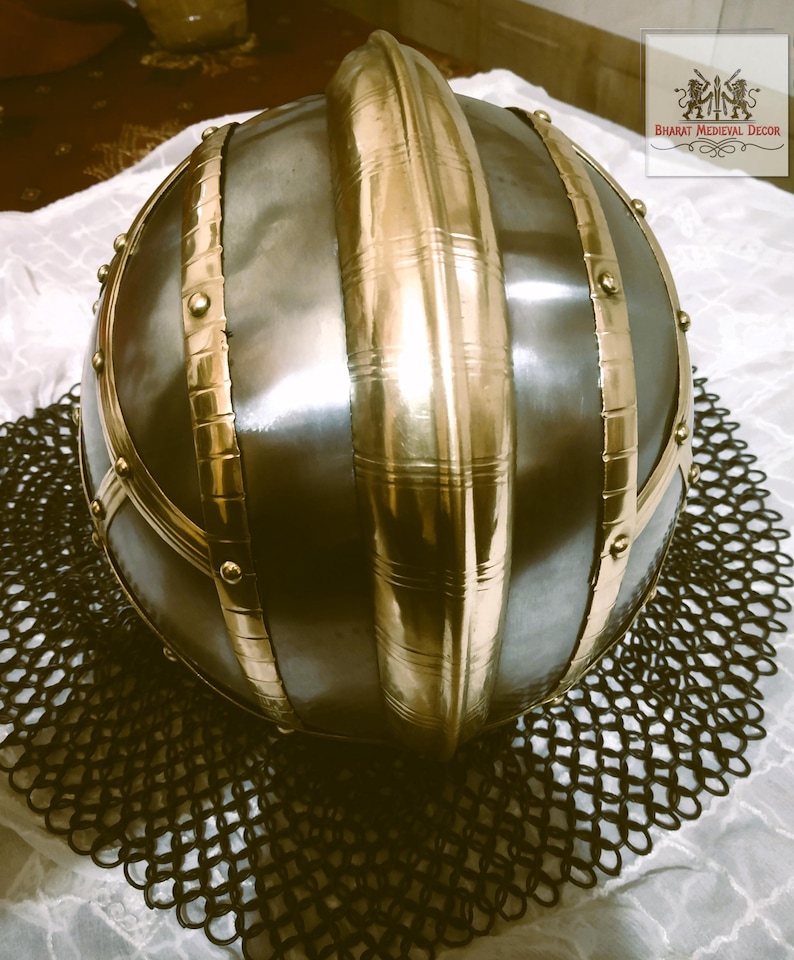 May include: A medieval-style helmet with a rounded, dark metal body and prominent gold-colored bands and accents. The helmet sits atop a chainmail-like mesh, suggesting historical armor or decorative item.