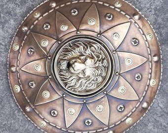 May include: A round, bronze-colored shield with a lion's head embossed in the center. The shield is decorated with a pattern of gold-colored studs and a rope-like border.