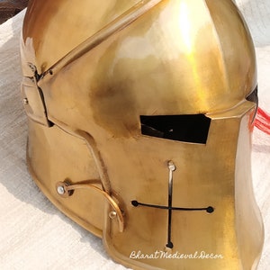 May include: A gold-colored medieval helmet with a cross design on the front. The helmet has a visor and is made of metal. The text "Bharat Medieval Decor" is visible on the top and bottom of the image.