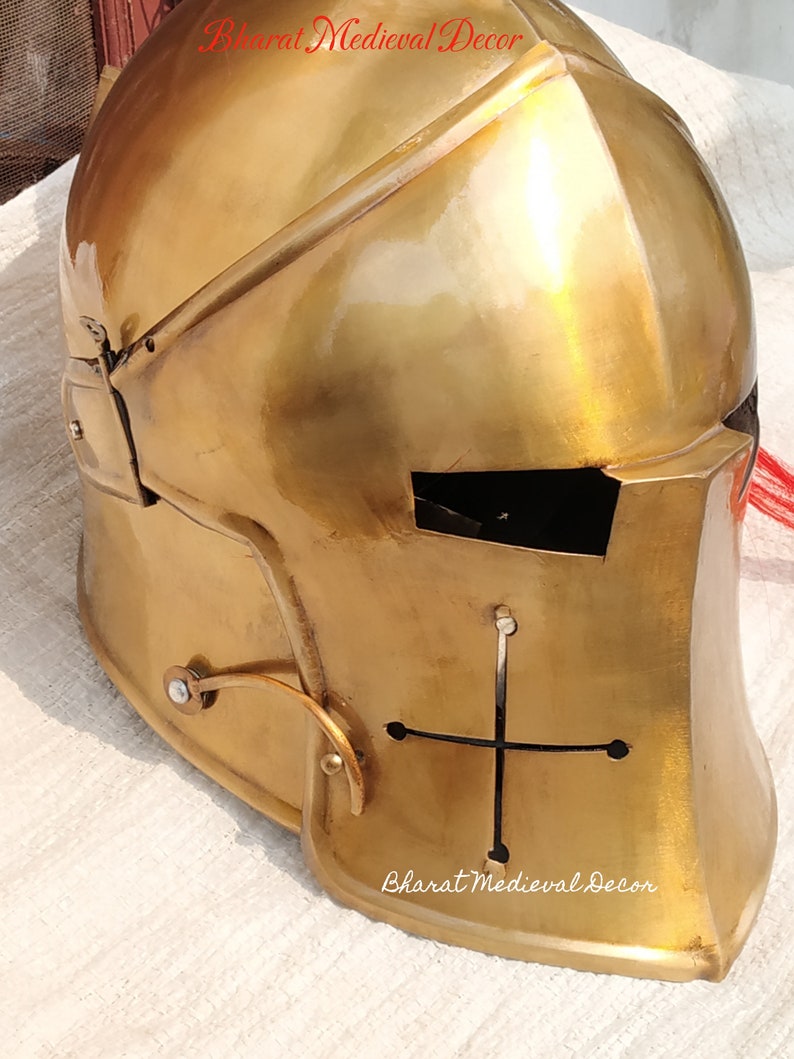May include: A gold-colored medieval helmet with a cross design on the front. The helmet has a visor and is made of metal. The text "Bharat Medieval Decor" is visible on the top and bottom of the image.