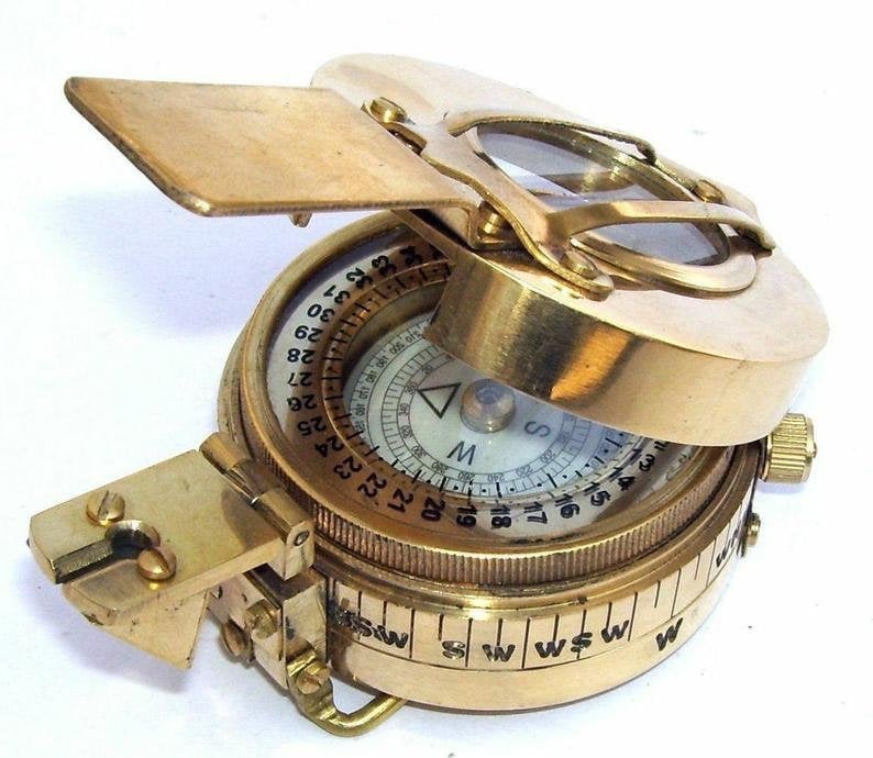 May include: A brass compass with a folding lid and a magnifying glass. The compass face has a white background with black markings and a red needle. The compass is marked with numbers from 1 to 360.