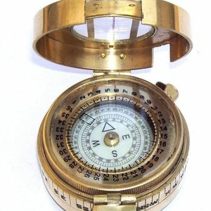 May include: A brass compass with a white face and black markings. The compass has a rotating bezel with numbers 1-36 and a compass rose with the letters N, S, E, and W. The compass is closed and has a hinged lid.