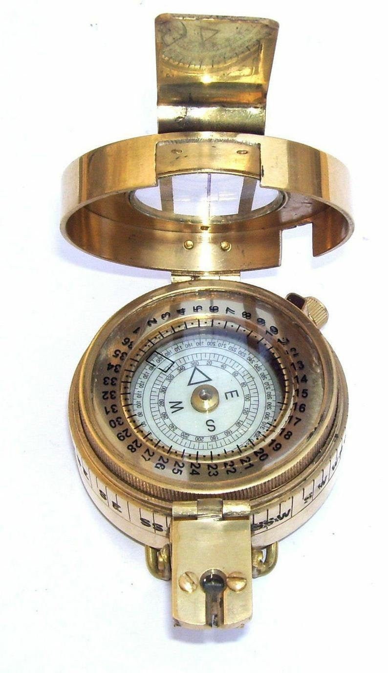 May include: A brass compass with a white face and black markings. The compass has a rotating bezel with numbers 1-36 and a compass rose with the letters N, S, E, and W. The compass is closed and has a hinged lid.
