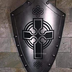 Puede incluir: Un escudo de metal plateado con un dise&ntilde;o de cruz celta negra. El escudo tiene una parte superior puntiaguda y una parte inferior redondeada. Est&aacute; decorado con remaches alrededor del borde.