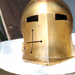 May include: A gold-colored medieval helmet with a cross design on the front. The helmet has two rectangular eye openings and a circular side detail. The text "Bharat Medieval Decor" is visible on the helmet.