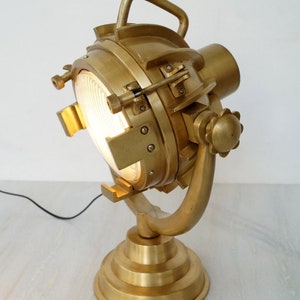 Può includere: Una lampada faro in ottone con una lente rotonda in vetro smerigliato e un braccio curvo. La lampada ha una base con un design a gradini e un cavo nero.