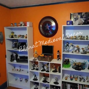 May include: White shelves filled with anime figurines and collectibles. The shelves are against an orange wall with a red, white, and blue Captain America shield on the wall. The text "Bharat Medieval Decor" is written on the wall.