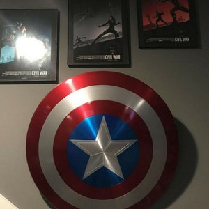 May include: A large, red, white, and blue Captain America shield with a silver five-pointed star in the center. The shield is hanging on a gray wall with three movie posters for Captain America: Civil War.