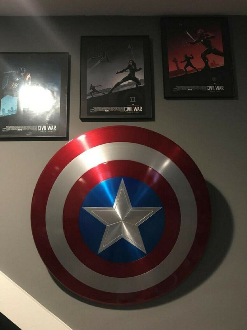 May include: A large, red, white, and blue Captain America shield with a silver five-pointed star in the center. The shield is hanging on a gray wall with three movie posters for Captain America: Civil War.