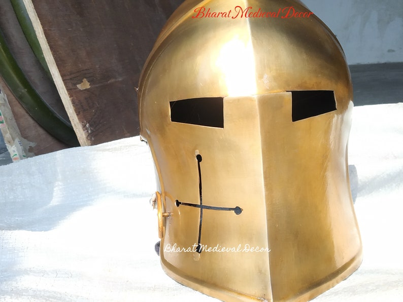 May include: A gold-colored medieval knight's helmet with a cross design. The helmet has eye slits and a smooth, reflective surface. The text "Bharat Medieval Decor" is visible on the helmet.