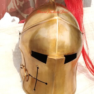 May include: A polished brass medieval helmet with a red plume. The helmet features a cross design on the front and a visor opening. The text "Bharat Medieval Decor" is visible on the side.