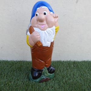 May include: A brightly coloured garden gnome statue. The gnome is depicted wearing a blue hat, white beard, and brown overalls. It holds a wooden tool and stands on a green base. The statue is painted with bright colours and has a cheerful expression.