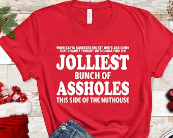 May include: A red t-shirt with white text that reads "When Santa squeezes his fat white ass down that chimney tonight, he's gonna find the Jolliest bunch of assholes this side of the nuthouse." The shirt is paired with blue jeans and black sneakers.