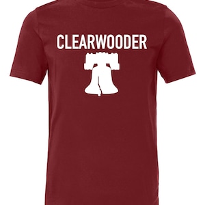 May include: A maroon t-shirt with the word "CLEARWOODER" in white block letters above a white silhouette of the Liberty Bell. The shirt is made of a soft material and has short sleeves.