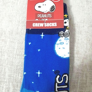 May include: Blue crew socks featuring Snoopy from Peanuts. The socks have a blue background with white stars and a large white moon. The packaging reads "Peanuts" and "Crew Socks."