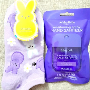 May include: A pair of lavender socks with a bunny and floral pattern, next to a yellow bunny-shaped tag. A purple hand sanitizer spray bottle is in a purple package, with the text "Ashley Belle" and "lavender drift".