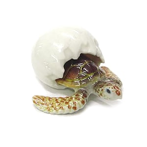May include: A ceramic figurine of a baby sea turtle emerging from a cracked white egg. The turtle has a brown and green shell, with a speckled pattern on its flippers and head. The figurine is set against a white background.