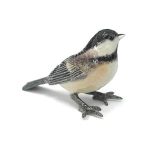 May include: A detailed figurine of a chickadee bird. The bird has a black head, white cheeks, and a tan body with gray wings. The bird is standing on two small feet, with its beak pointing upwards.