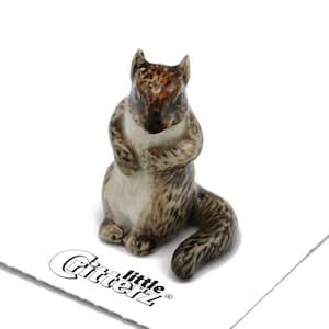 May include: A small ceramic squirrel figurine in shades of brown, tan, and white. The squirrel is in a seated position with its paws clasped. The figurine has a glossy finish and is set against a white background. The logo "Gitterz little" is visible.