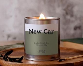 May include: A burning candle in a silver tin, labelled "New Car". The label details the scent as leather, linen, and ylang, with a 30-hour burn time and 250g of wax. The candle rests on a wooden tray, alongside a sprig of foliage and a used match.