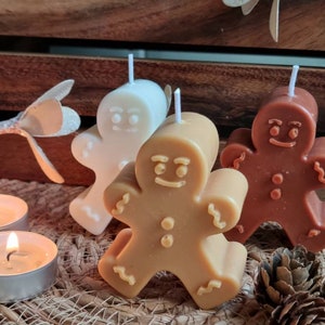 May include: Three gingerbread man shaped candles, one white, one brown, and one tan. The candles are sitting on a woven basket surface.