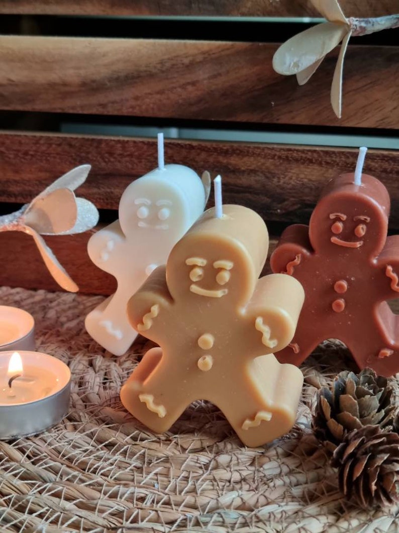 May include: Three gingerbread man shaped candles, one white, one brown, and one tan. The candles are sitting on a woven basket surface.