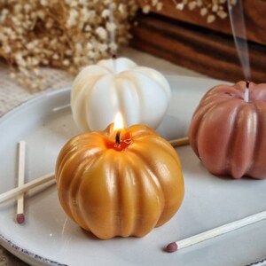 May include: Three pumpkin-shaped candles on a white plate. One candle is lit and is a golden yellow color. The other two candles are unlit and are white and a light brown color.