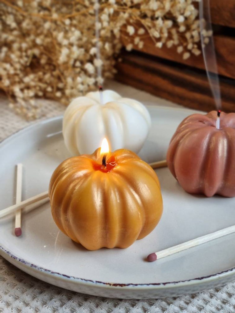 May include: Three pumpkin-shaped candles on a white plate. One candle is lit and is a golden yellow color. The other two candles are unlit and are white and a light brown color.
