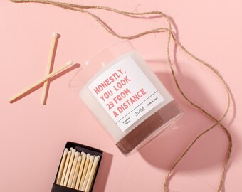 May include: A clear glass candle with a white label featuring the text "HONESTLY, YOU LOOK 29 FROM A DISTANCE." and "Pumpkin Spice". The candle is accompanied by a small black box of wooden matches and two loose matches on a pink surface.