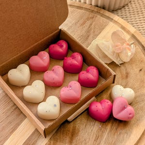 May include: A box of heart-shaped wax melts in pink, white, and red colors. The melts are arranged in a box with a lid. The box is made of brown cardboard.