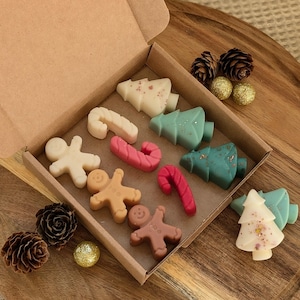 May include: A box of Christmas-themed wax melts in various shapes and colors, including gingerbread people, candy canes, and Christmas trees. The melts are arranged in a brown cardboard box.