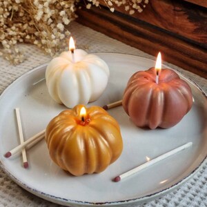 May include: Three pumpkin-shaped candles on a white plate. The candles are lit and are in the colors white, orange, and brown. There are also matches on the plate.