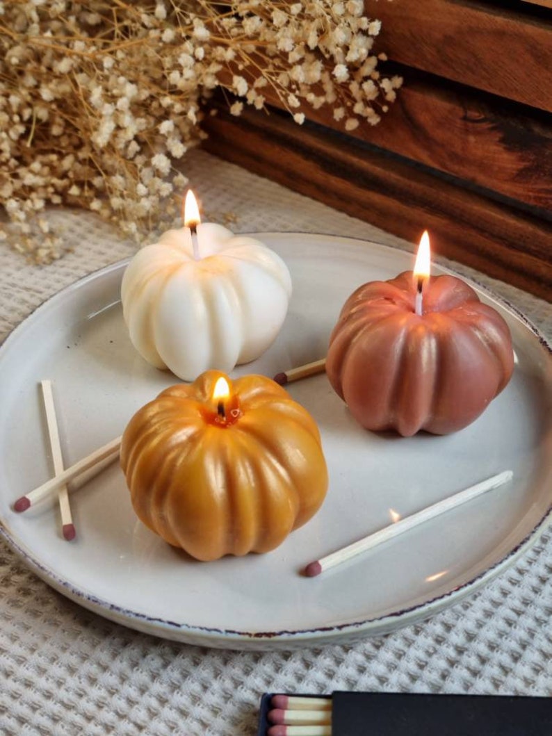 May include: Three pumpkin-shaped candles on a white plate. The candles are lit and are in the colors white, orange, and brown. There are also matches on the plate.
