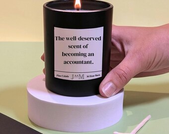 May include: Black candle with a white label that reads "The well-deserved scent of becoming an accountant." The candle is labeled "Pina Colada" and "JAM" and has a 30-hour burn time.