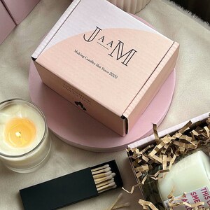 以下が含まれることがあります： 前面に「JAM Making Candles Hot Since 2020」と書かれたピンクの箱。箱はピンクの表面の上に置かれています。ガラスの容器に入った白いろうそくとマッチの箱が表面にあります。