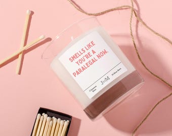 May include: A clear glass candle with a white label that reads "Smells like you're a paralegal now." The candle has a brown base and is next to a box of matches and two crossed matches. The candle is labeled "Cinnamon Buns" and has a 30-hour burn time.
