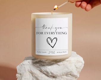 May include: A white candle with a label that reads "thank you for everything" and a heart symbol. The candle is on a white stone. A lit match is held above the candle wick. The candle has a 40-hour burn time.