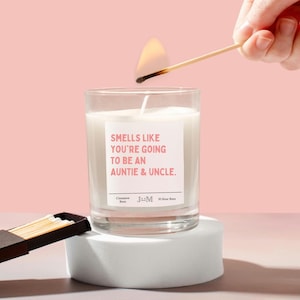 May include: A white candle with a pink label that reads "Smells like you're going to be an auntie & uncle." The candle is on a white pedestal and a hand is lighting the candle with a match.