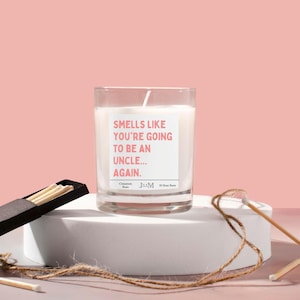Può includere: Una candela bianca in un barattolo di vetro trasparente con un'etichetta rosa che dice "Smells like you're going to be an uncle... again." La candela è su una superficie bianca con una scatola di fiammiferi e uno spago.
