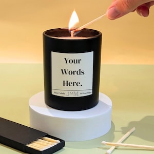 May include: A burning black candle with a white label reading "Your Words Here." The candle is on a white stand, with a box of matches and loose matches. The candle is labelled "Pina Colada" and "30 Hour Burn."