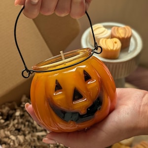K&ouml;nnte beinhalten: Orangefarbene K&uuml;rbiskerze mit schwarzem Griff und geschnitztem Jack-o'-Lantern-Gesicht. Die Kerze hat einen Docht und wird in einer Hand gehalten. Weitere k&uuml;rbisf&ouml;rmige Kerzen befinden sich im Hintergrund.