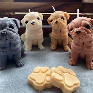 Puede incluir: Cuatro velas en forma de perro hechas a mano. Las velas son de color gris, blanco, marrón y beige. Las velas están sobre una superficie de tela azul claro.