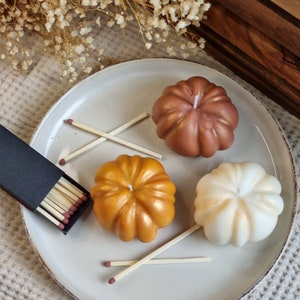May include: Three pumpkin-shaped candles on a white plate. The candles are in shades of brown, gold, and white. There are also matches in a black box on the plate.