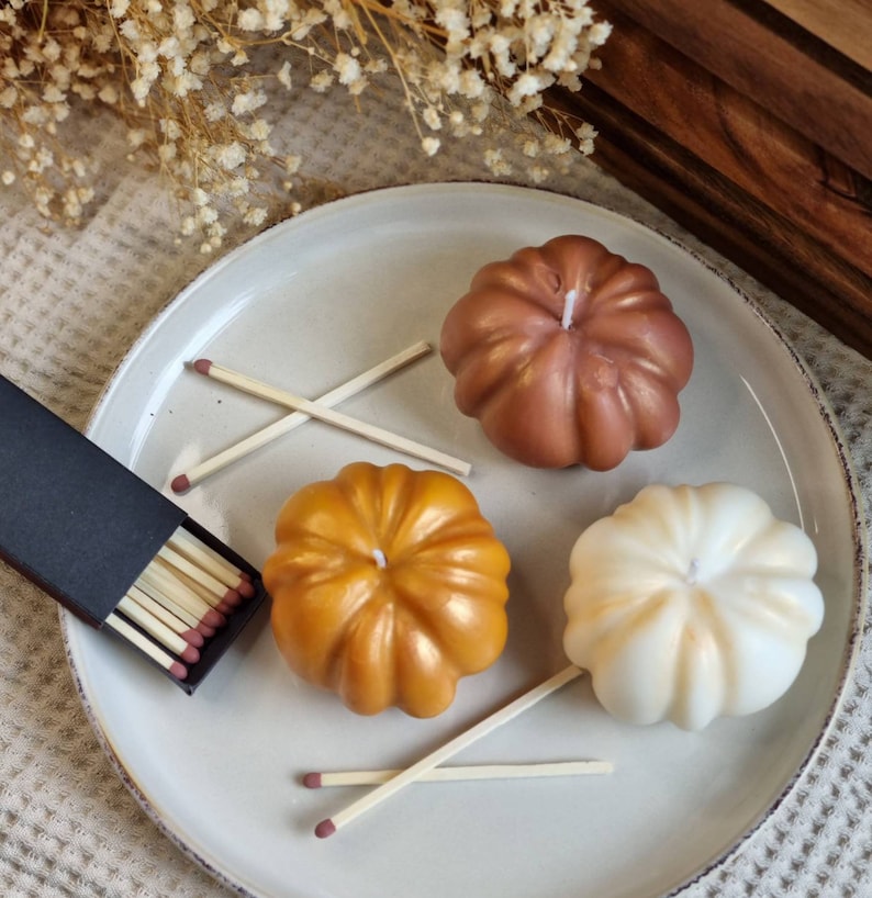May include: Three pumpkin-shaped candles on a white plate. The candles are in shades of brown, gold, and white. There are also matches in a black box on the plate.
