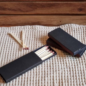 May include: Two black matchboxes with a stack of wooden matches in one of the boxes. The matches are on a beige textured surface.