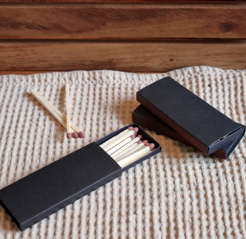 May include: Two black matchboxes with a stack of wooden matches in one of the boxes. The matches are on a beige textured surface.